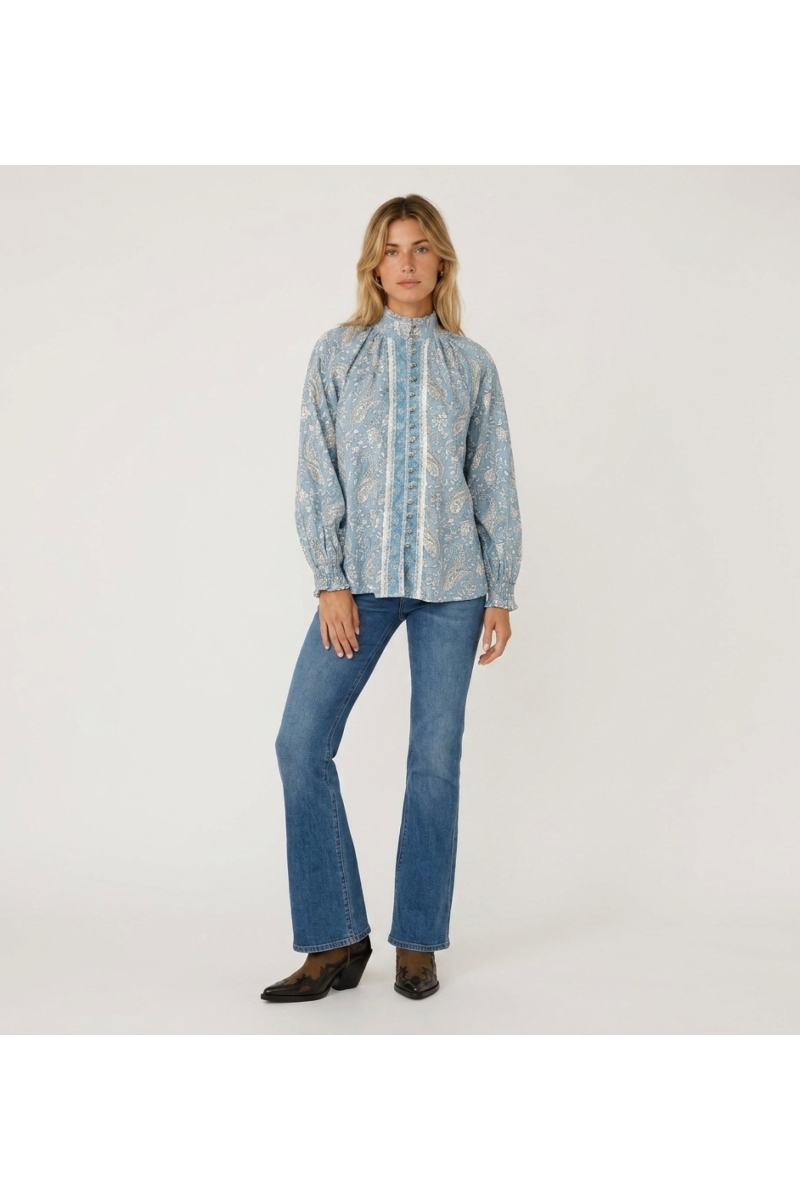 Blusa azul claro de manga larga con estampado cachemira blanco, cuello alto, abotonada al frente y puños con volantes.