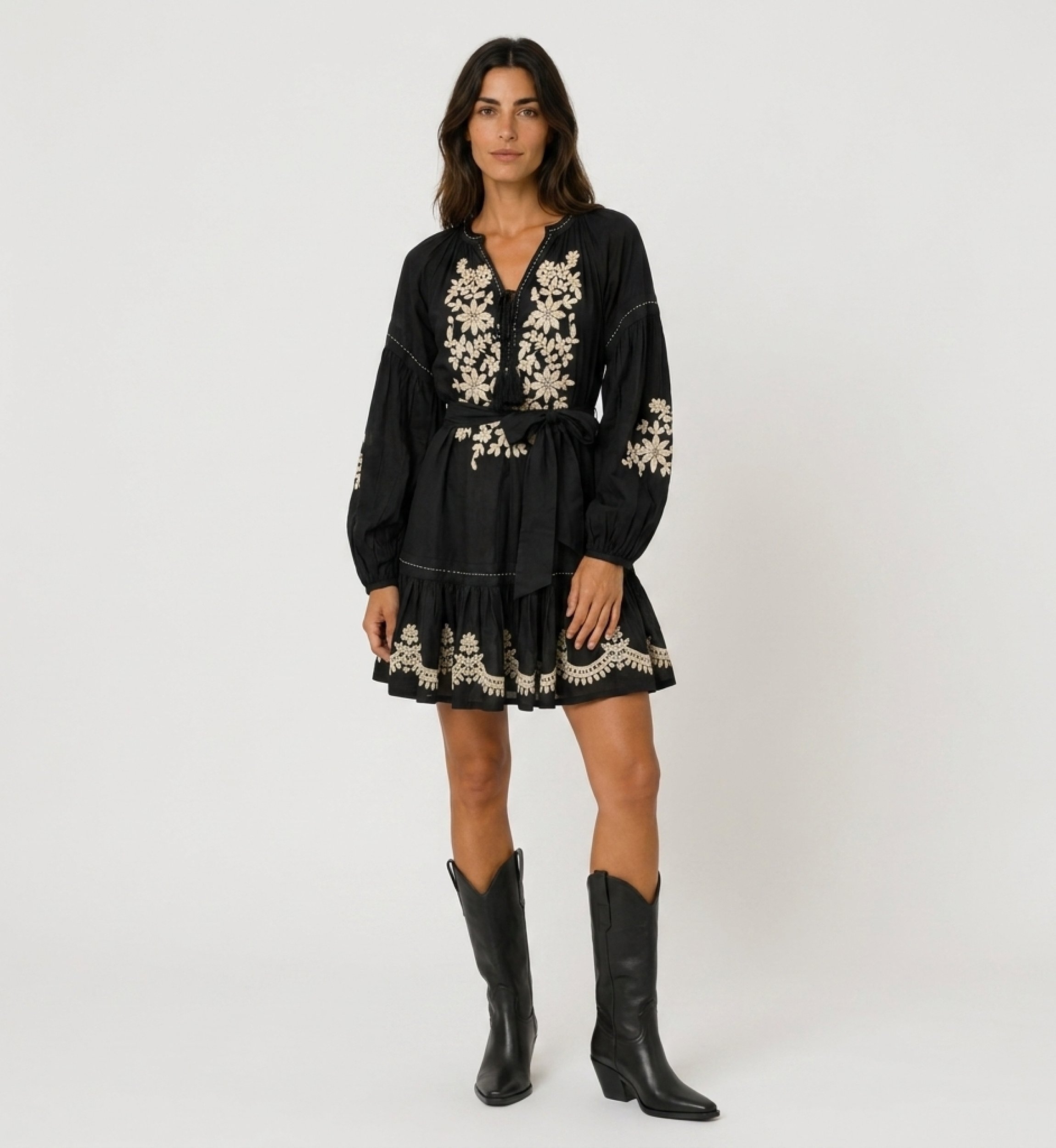 Mini-robe noire ample à manches longues ballon, jupe étagée, broderie florale blanche et taille à nouer, vue de face.