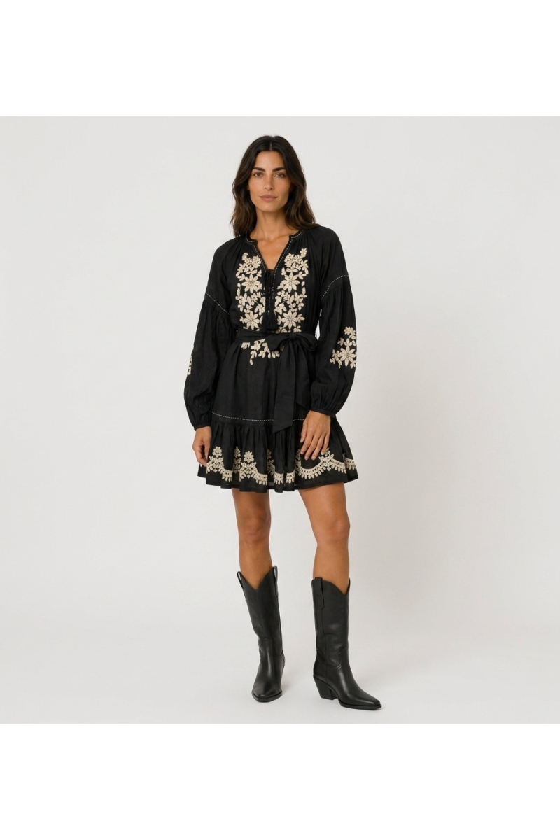 Mini-robe noire ample à manches longues ballon, jupe étagée, broderie florale blanche et taille à nouer, vue de face.
